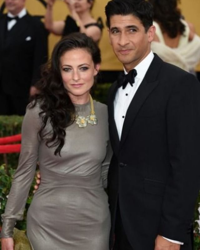 Raza Jaffrey in black formal dress posing with his wife Lara Pulver in a grey bodycon and golden jewelry.